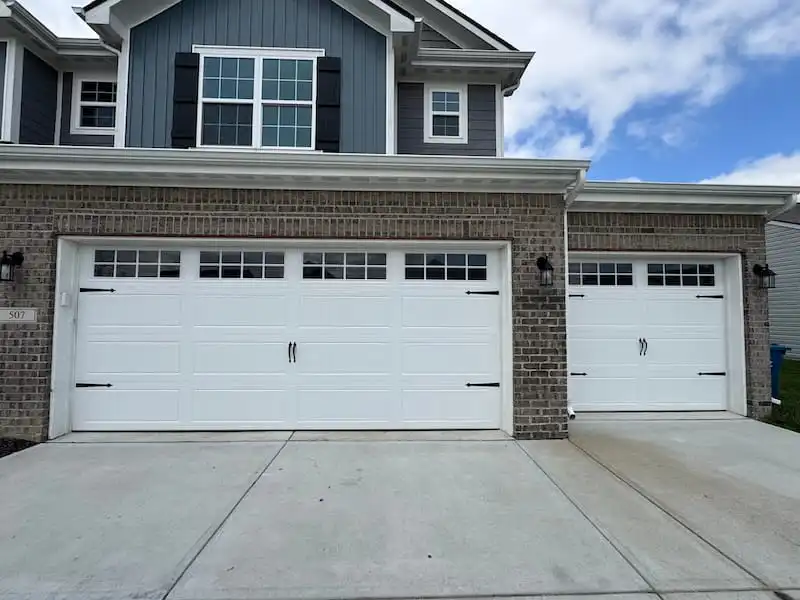 Two white carriage style garage doors with black decorative hardware and upper windows installed on a two-car and single-car garage of a brick home in Carmel, IN